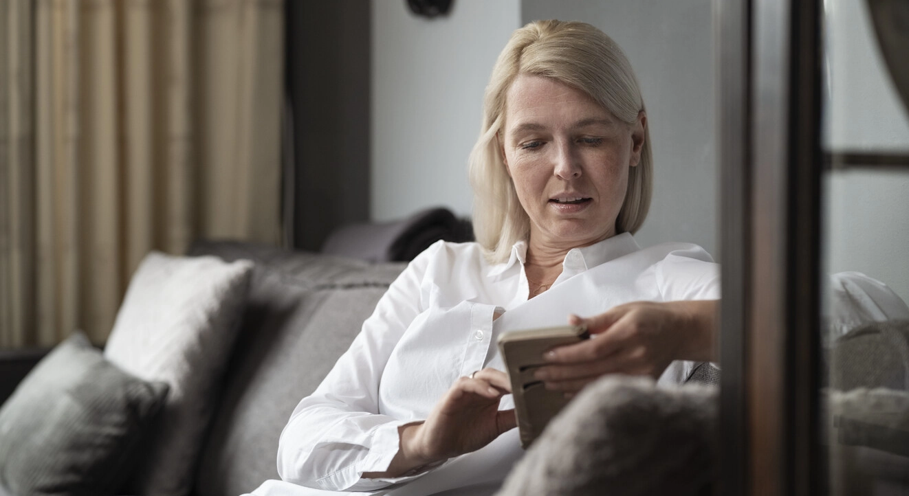 Bild von Ilse, die in ihrem Wohnzimmer sitzt. Multiple Sklerose schädigt das Nervensystem, stört die Signalübertragung von Gehirn und Rückenmark und kann daher eine Vielzahl von Körperfunktionen beeinträchtigen. Ihre Blase kann davon betroffen sein.  